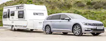 Silberner Kombi-PKW zieht weißen Wohnwagen mit EN-Wohnmobil-Beschriftung, geparkt auf einem Schotterplatz vor grüner Böschung.