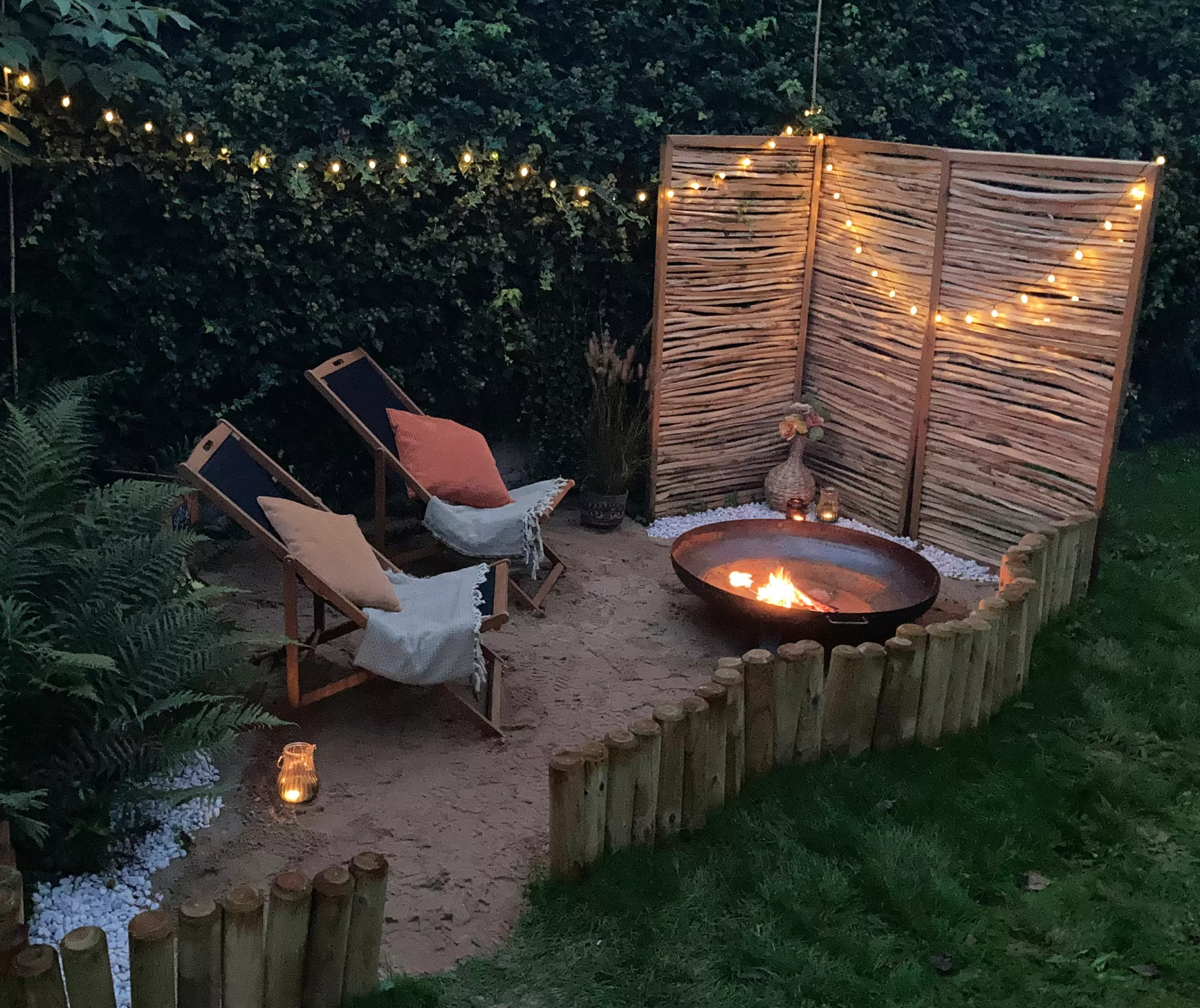 Ein Strand im Garten, darauf 2 Liegestühle, und eine Feuerschale. Man sieht eine Lichterkette und ein Sichtschutz.