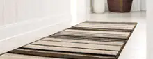 Läufer im Flur mit Streifenmuster in Beige- und Brauntönen auf hellem Boden, daneben weiße Wand und Pflanzgefäß.