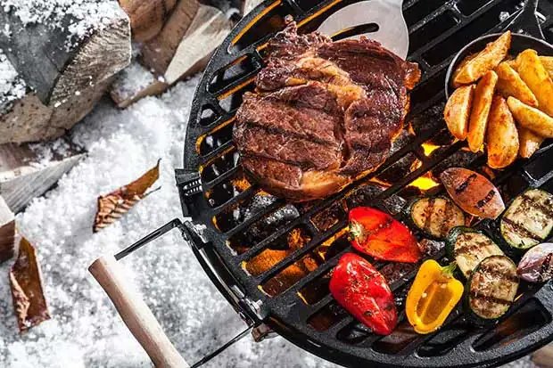 Steak, Kartoffelspalten und buntes Grillgemüse auf einem heißen Grill im Schnee, mit brennender Glut und Holzscheiten im Hintergrund.