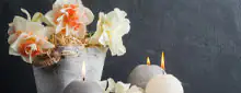 Dekoratives Arrangement mit weißen und grauen runden Kerzen, weißen Blumen und einem grauen Topf vor dunklem Hintergrund.