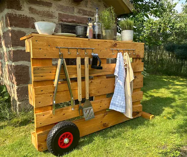 Die Grillstation aus Paletten steht auf einer Wiese. Es gibt genügend Platz, um Grillutensilien, Kräuter und Küchenhandtücher aufzubewahren-