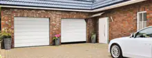 Moderne Hausfassade mit zwei weißen Sektionaltoren, rotem Klinker, Haustür und geparktem weißen Auto auf gepflastertem Hof.